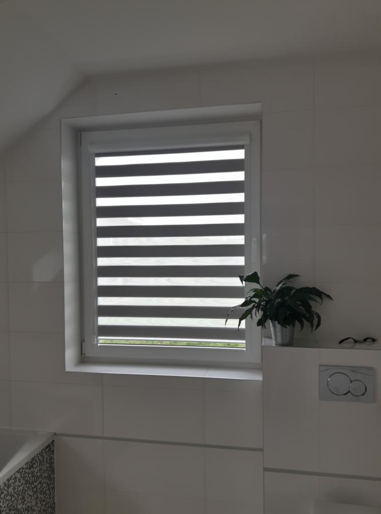 Okno w łazience na poddaszu z zamontowaną roletą wewnętrzną typu dzień i noc, obok doniczka z zieloną rośliną.