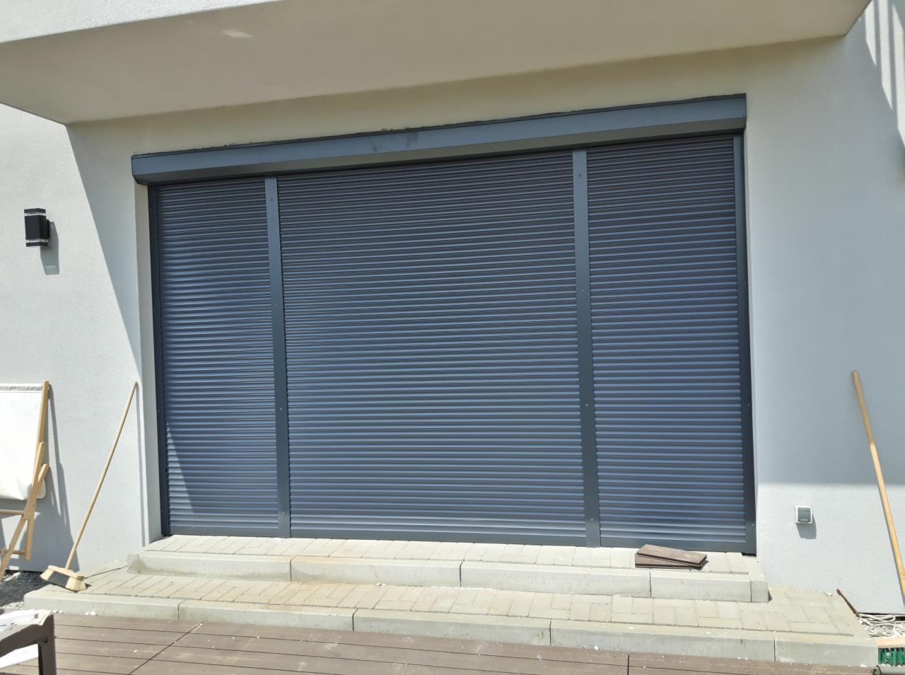 Zamknięta, grafitowa osłona okienna na parterze domu z widocznym fragmentem tarasu i białej ściany.