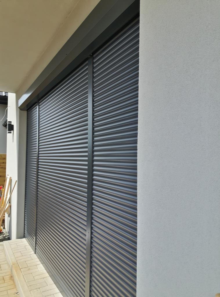 Zewnętrzna, antracytowa osłona okienna z lamelami, zamontowana na elewacji budynku z jasnoszarym tynkiem strukturalnym.