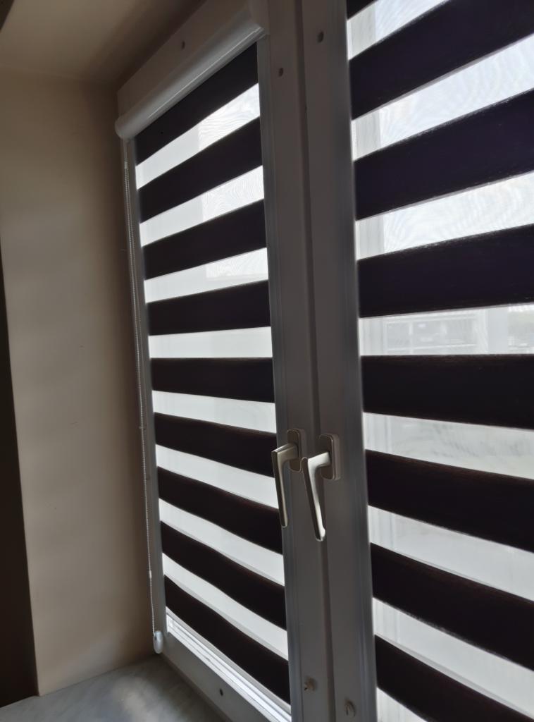 Okno z zamontowaną wewnętrzną przesłoną okienną typu dzień i noc, z widocznym mechanizmem sterującym i poziomymi pasami w kolorach bieli i brązu.