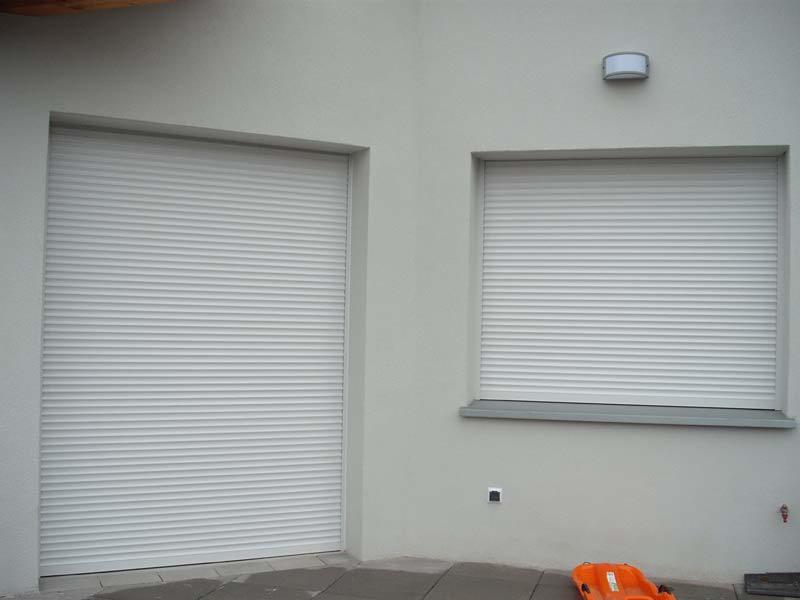 Dwa okna z zewnętrznymi osłonami przeciwsłonecznymi, jedno większe, drugie mniejsze, na szarej ścianie budynku, z pomarańczowym sankami na ziemi.