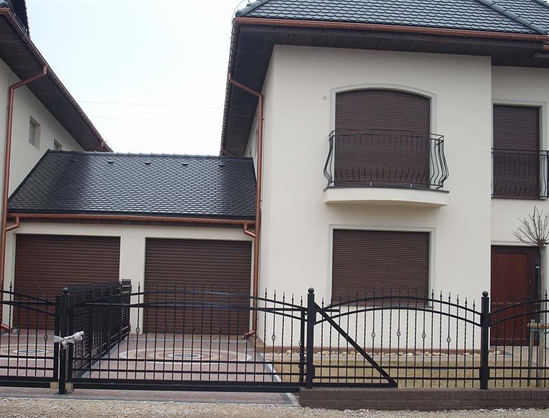 Dwukondygnacyjny dom z brązowymi roletami zewnętrznymi na oknach i bramie garażowej, czarne metalowe ogrodzenie z przesuwną bramą wjazdową.