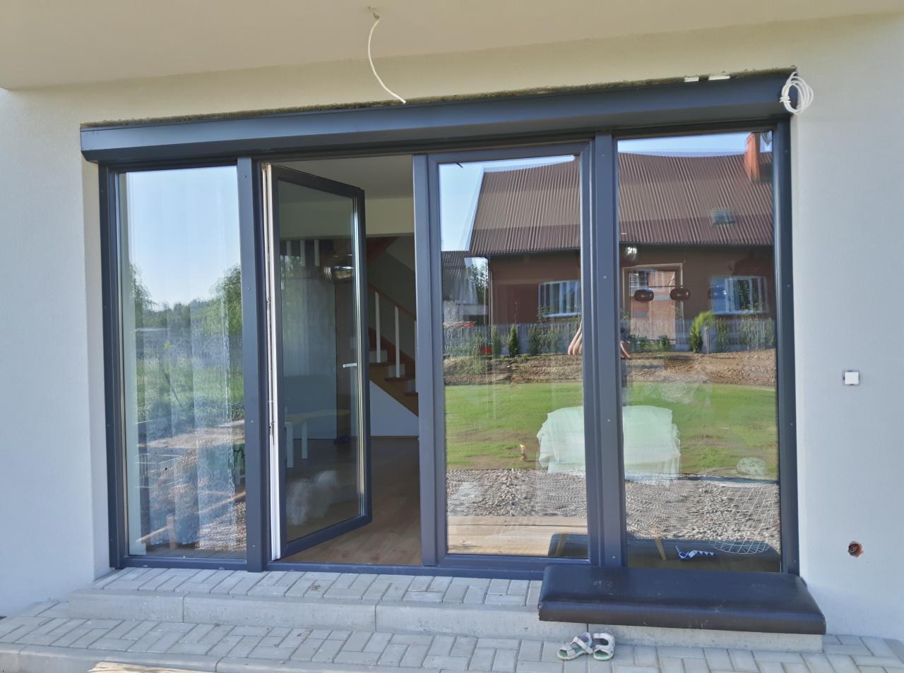 Szare, przeszklone drzwi tarasowe z zewnętrzną osłoną zwiniętą u góry, widok na ogród i dom, częściowo otwarte drzwi do wnętrza.