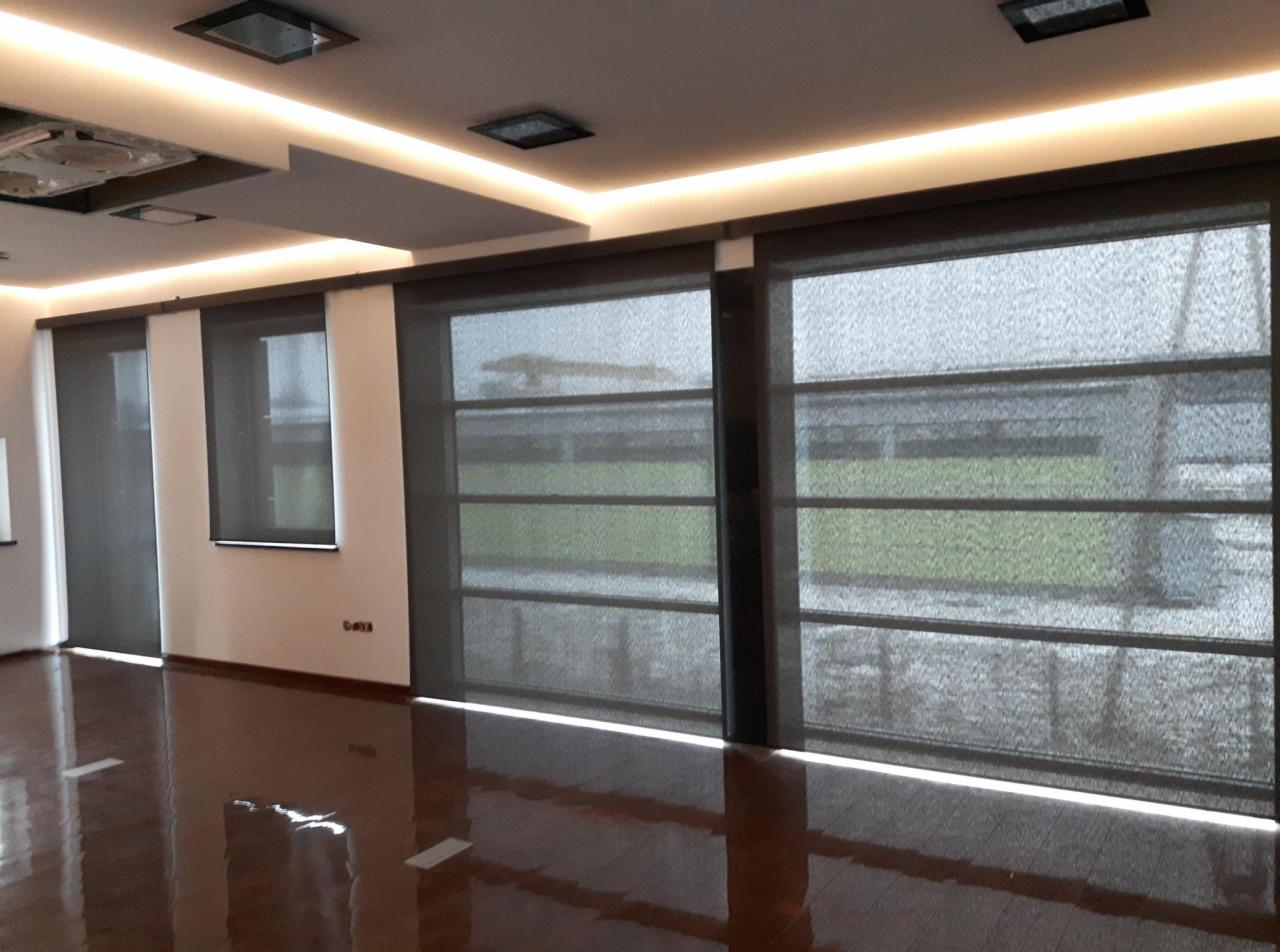 Nowoczesne, szare przesłony okienne w przestronnym salonie z błyszczącą podłogą, oświetleniem sufitowym LED i widokiem na zieleń za oknem.