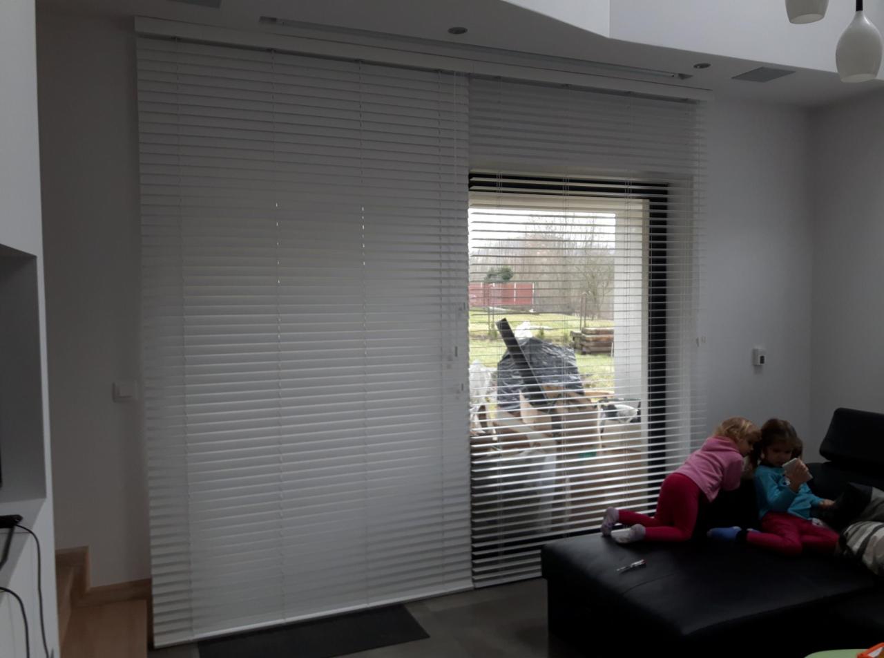 Salon z dużym oknem przesłoniętym białymi żaluzjami poziomymi; dwie dziewczynki bawią się na kanapie w tle.