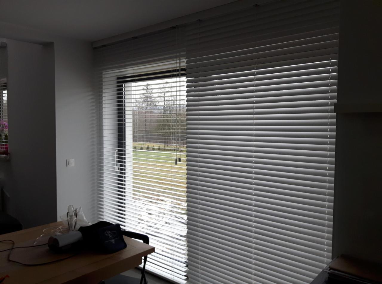 Białe poziome osłony okienne zamontowane w oknie z widokiem na zewnątrz, częściowo zasłonięte.