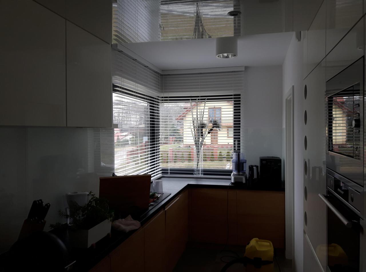 Kuchnia z nowoczesnymi, białymi żaluzjami poziomymi w oknach narożnych, widok na dom z ogrodem, odblaskowy sufit.
