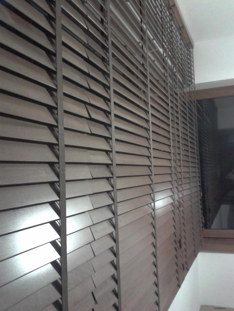 Ciemnobrązowe, drewniane lamele okienne, częściowo otwarte, widoczne z boku, z oknem w tle.