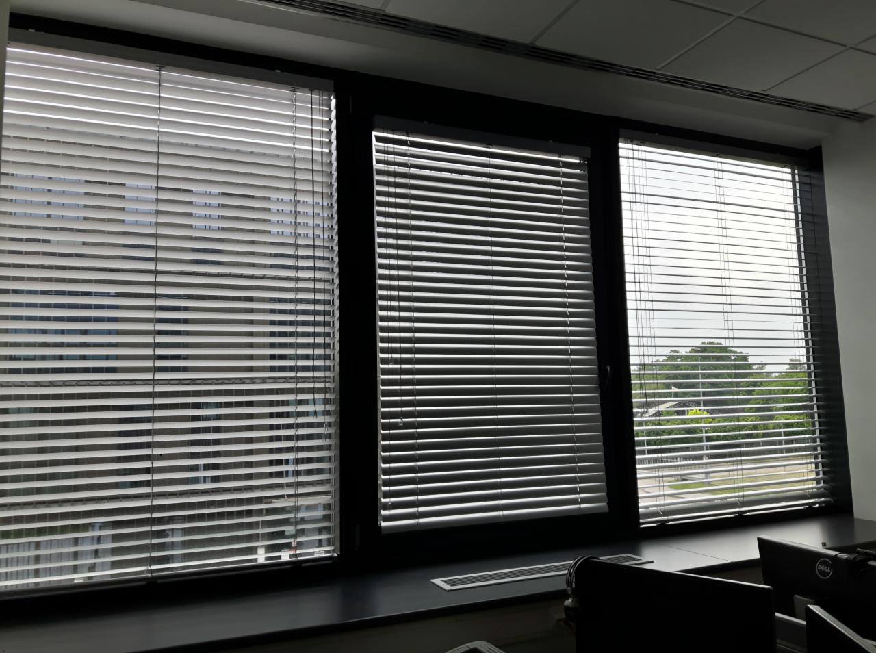 Trzy okna z poziomymi listwami przeciwsłonecznymi w biurze, widok na budynek i zieleń na zewnątrz.