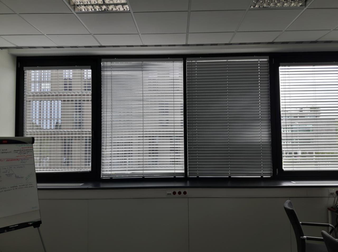 Biuro z trzema oknami wyposażonymi w poziome żaluzje aluminiowe w odcieniach szarości, z widocznym fragmentem tablicy suchościeralnej po lewej stronie.
