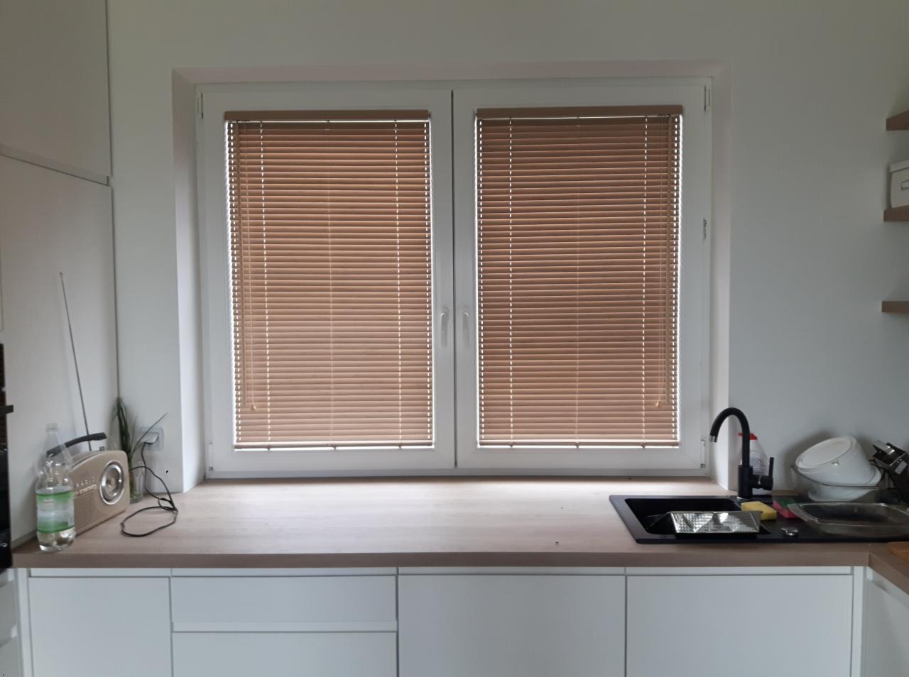 Brązowe listkowe osłony okienne w białej ramie okiennej, widok z wnętrza kuchni, na blacie kuchennym radio i naczynia w zlewie.