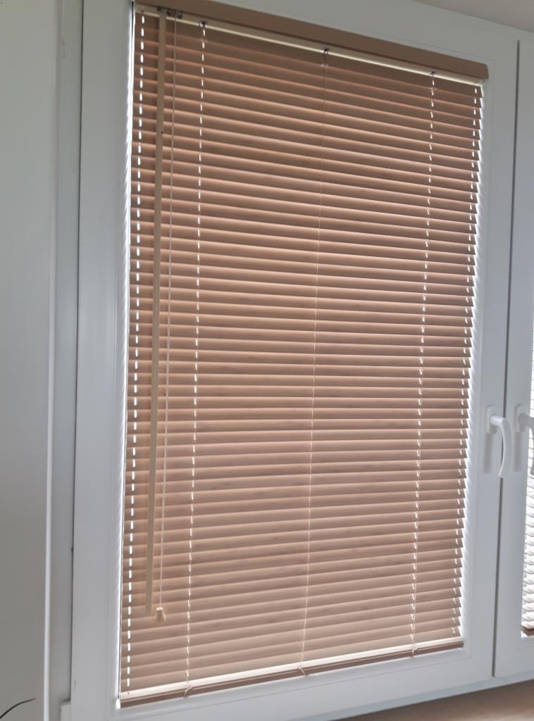Brązowe poziome osłony okienne zamontowane w białej ramie okiennej, widoczne sznurki sterujące.