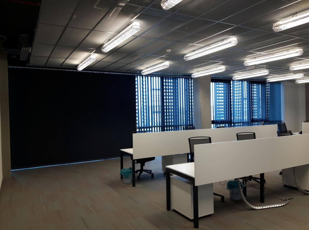 Biuro z pionowymi osłonami okiennymi w kolorze niebieskim i czarnym, widoczne biurka z białymi panelami działowymi i krzesła biurowe.