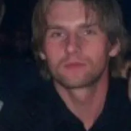 Portret blond mężczyzny w czarnej koszuli, pozującego w słabym oświetleniu.