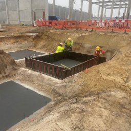 Brother Groups Sp. z o.o. - Pracownicy w żółtych kamizelkach pracują przy szalunku zalanym betonem w wykopie, widoczne stalowe elementy konstrukcyjne budynku w tle.