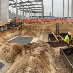 Brother Groups Sp. z o.o. - Szeroki kadr na plac budowy z wykopami pod fundamenty, częściowo zalanymi betonem, z widocznymi pracownikami w odblaskowych kamizelkach, zbrojeniami i szalunkami, na tle konstrukcji stalowej...