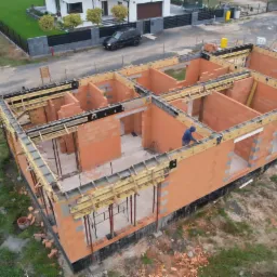 Widok z góry na budowę domu jednorodzinnego z czerwonej cegły, z widocznym szalunkiem na wieńcu i pracownikiem w niebieskiej bluzie. Na działce stoi przenośna toaleta, a w tle zaparkowany samochód...