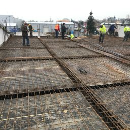 Brother Groups Sp. z o.o. - Zbrojenie płyty fundamentowej z widocznymi pracownikami budowlanymi w tle, przygotowane pod wylanie betonu, z widocznymi deskami szalunkowymi.