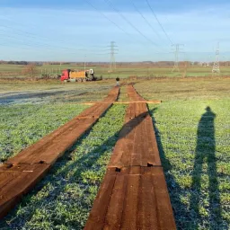 Tymczasowa droga z drewnianych belek ułożona na zroszonej trawie, z widocznym pracownikiem i ciężarówką w tle, pod liniami wysokiego napięcia.