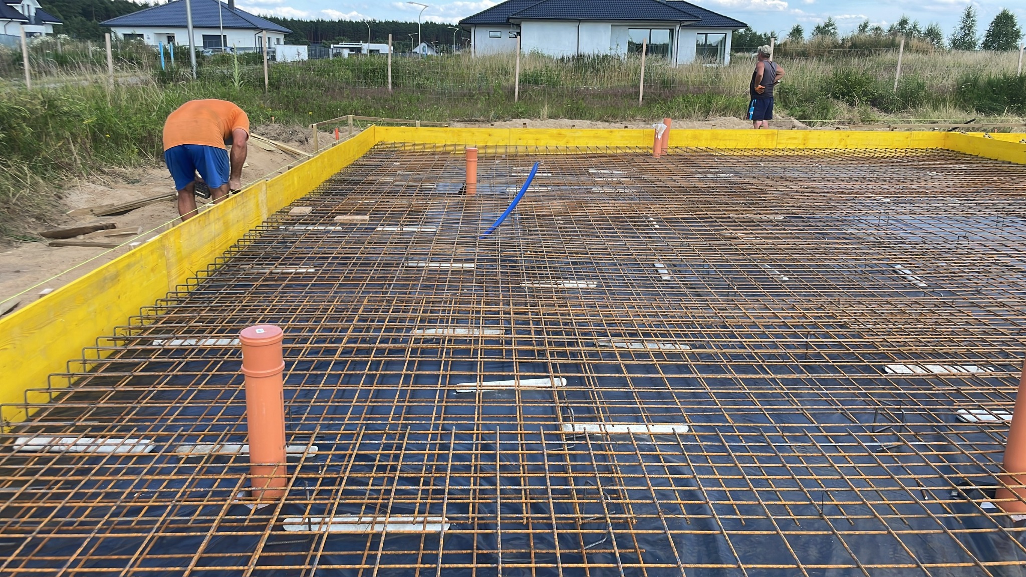 Przygotowanie zbrojenia fundamentów pod budowę domu jednorodzinnego, widoczna siatka z prętów zbrojeniowych, szalunek z żółtych desek oraz wystające rury instalacyjne.