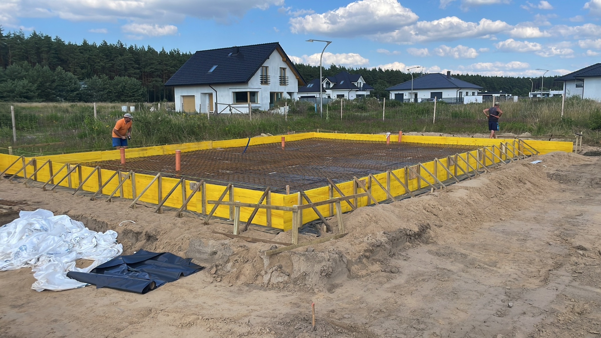 Przygotowanie zbrojenia fundamentów pod dom jednorodzinny z żółtym szalunkiem, widoczni pracownicy budowlani i okoliczne domy.