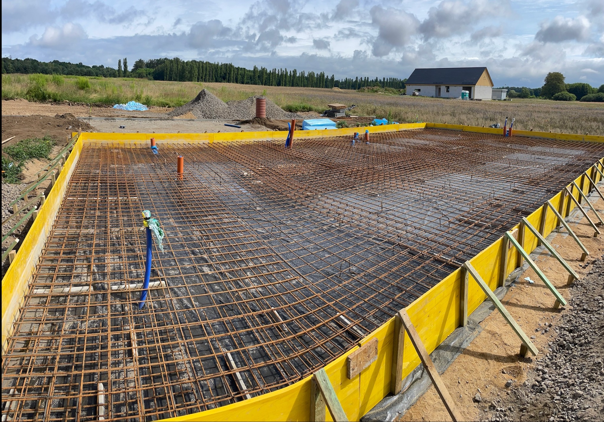 Przygotowanie zbrojenia pod płytę fundamentową z żółtym szalunkiem, widoczne pionowe rury instalacyjne oraz dom w tle.