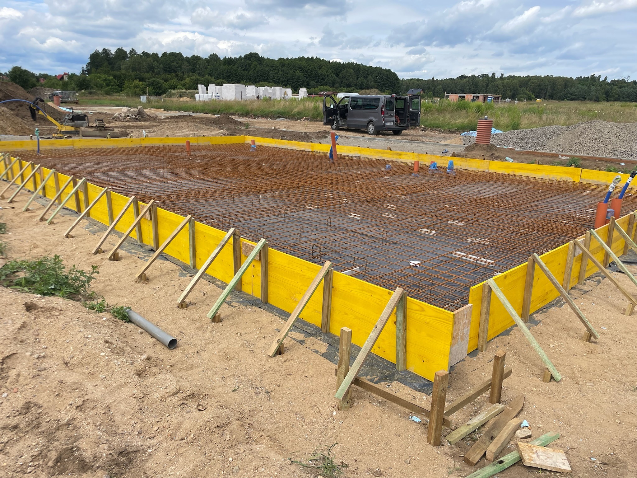Przygotowanie zbrojenia i szalunku z żółtych płyt na płycie fundamentowej pod budowę domu jednorodzinnego, widoczny szary samochód dostawczy z otwartymi drzwiami i koparka w tle.