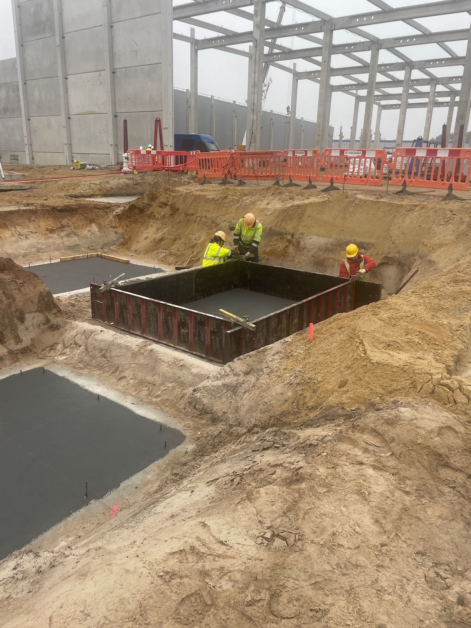 Pracownicy w żółtych kamizelkach pracują przy szalunku zalanym betonem w wykopie, widoczne stalowe elementy konstrukcyjne budynku w tle.