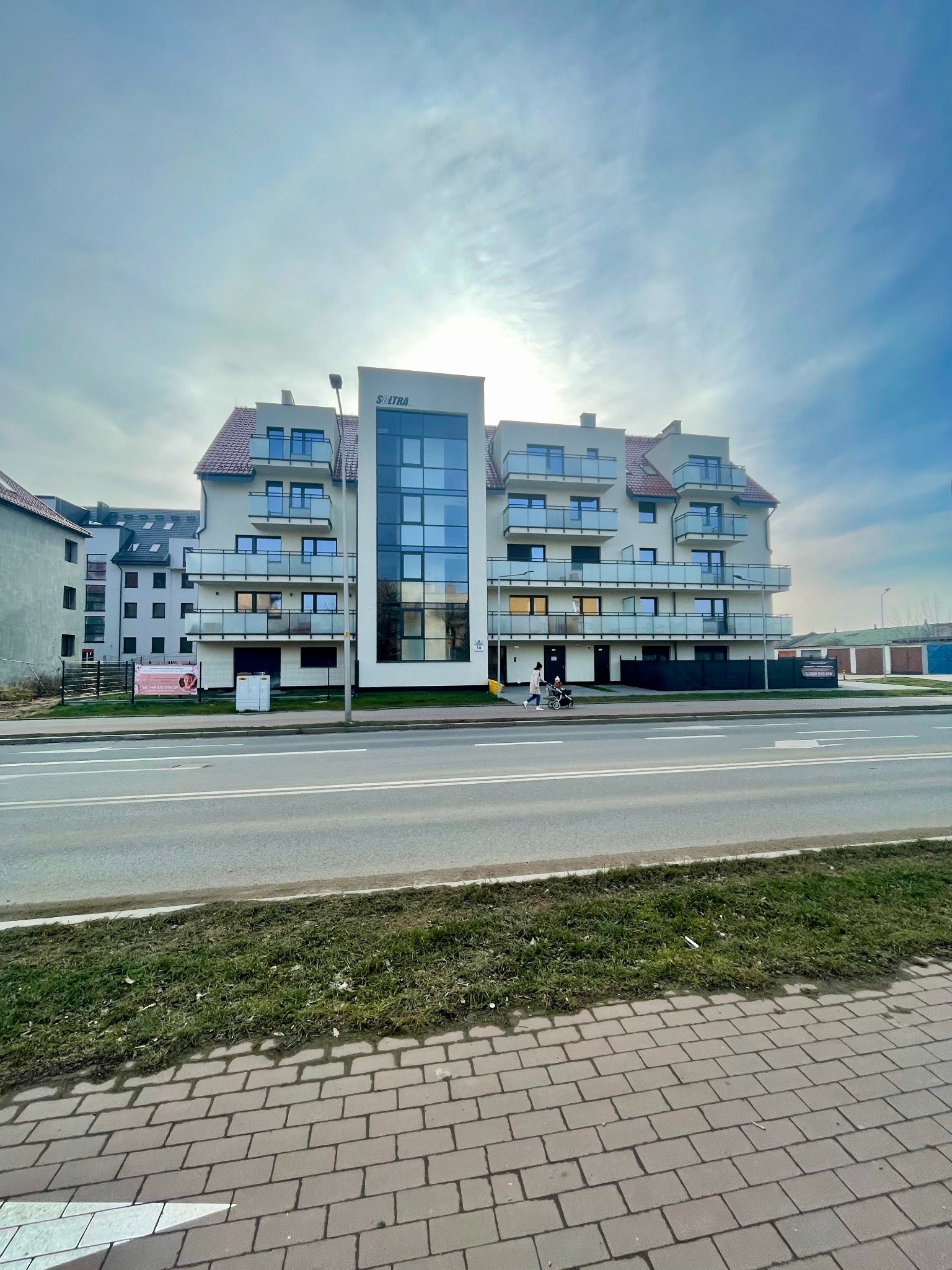 Nowoczesny budynek mieszkalny z balkonami o szklanych balustradach, widoczny z perspektywy ulicy, z reklamą na ogrodzeniu i osobą z wózkiem na chodniku.