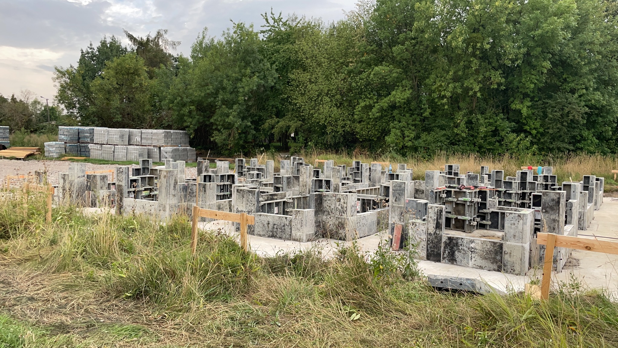 Wznoszenie fundamentów domu: betonowe szalunki ustawione na przygotowanym terenie, w tle palety z materiałami budowlanymi i zieleń drzew.