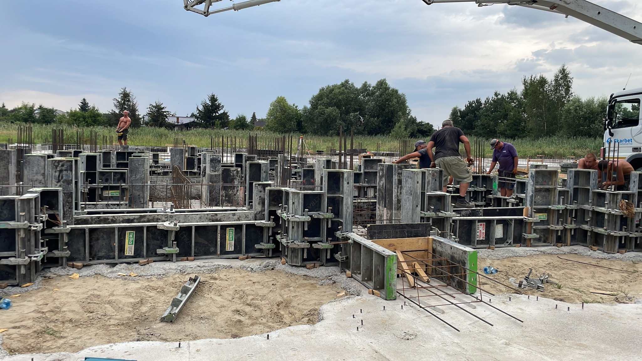 Montaż szalunków fundamentowych z widocznym zbrojeniem i pracownikami budowlanymi, w tle pompa do betonu i zieleń