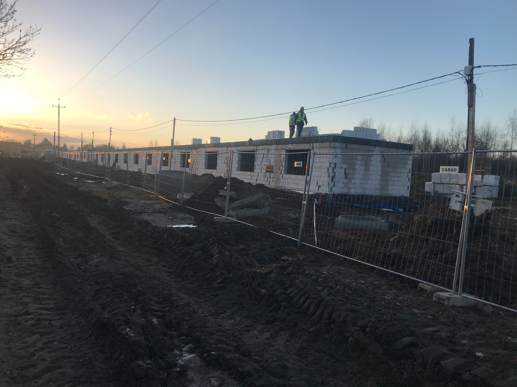 Długi szereg budynków w trakcie wznoszenia ścian z białych bloczków, widoczni dwaj pracownicy w odblaskowych kamizelkach na dachu, teren budowy ogrodzony siatką, widoczne elementy instalacji...