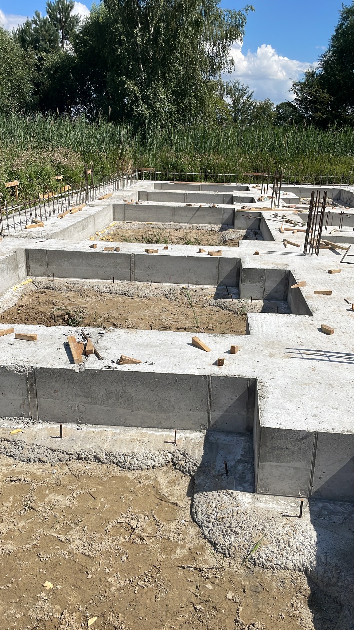 Świeżo wylane betonowe fundamenty domu jednorodzinnego z wystającymi prętami zbrojeniowymi, widok z góry na tle zieleni.