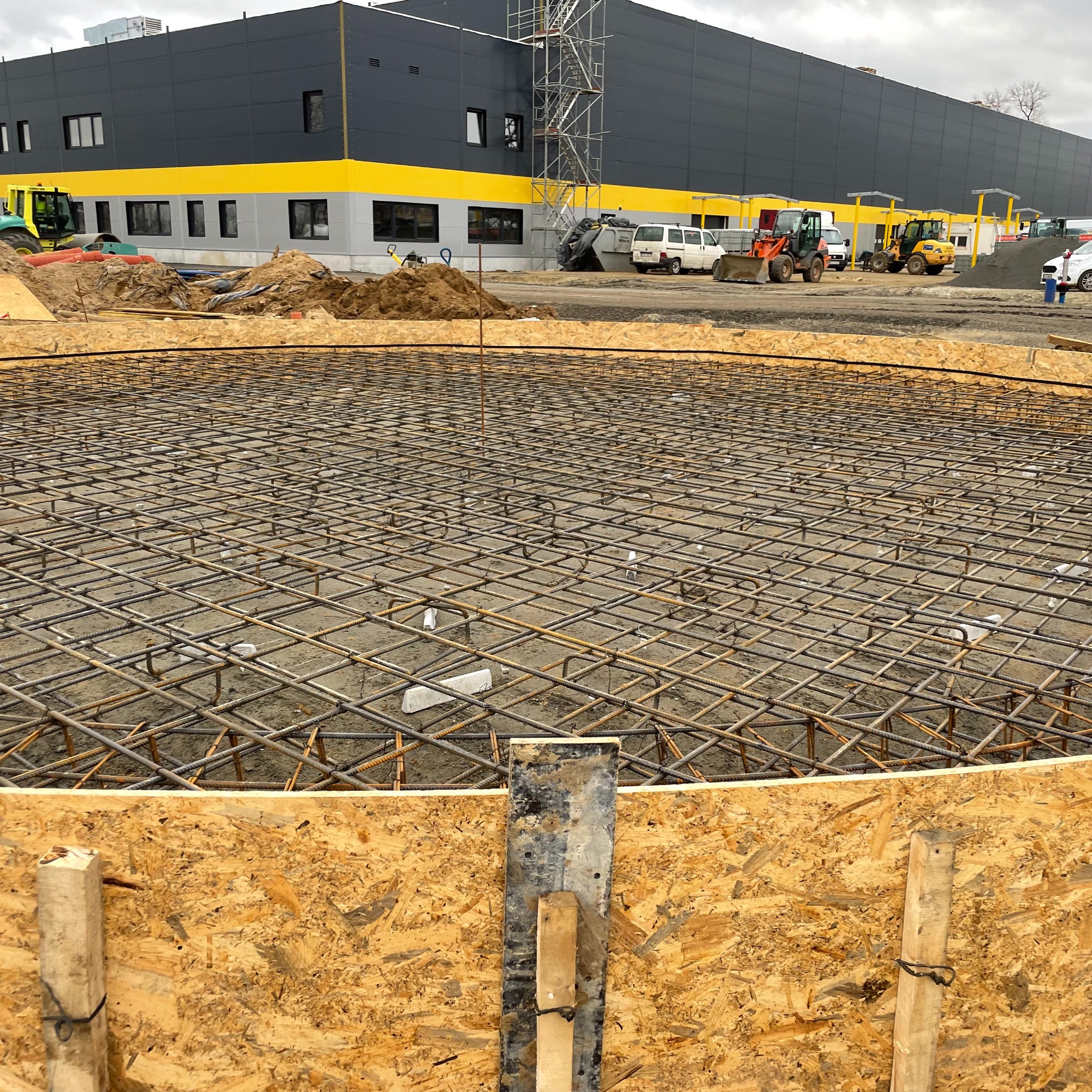 Zbrojenie okrągłego fundamentu pod budynek przemysłowy, widoczne szalunki z płyty OSB, w tle maszyny budowlane i hala w kolorach szaro-żółtych.