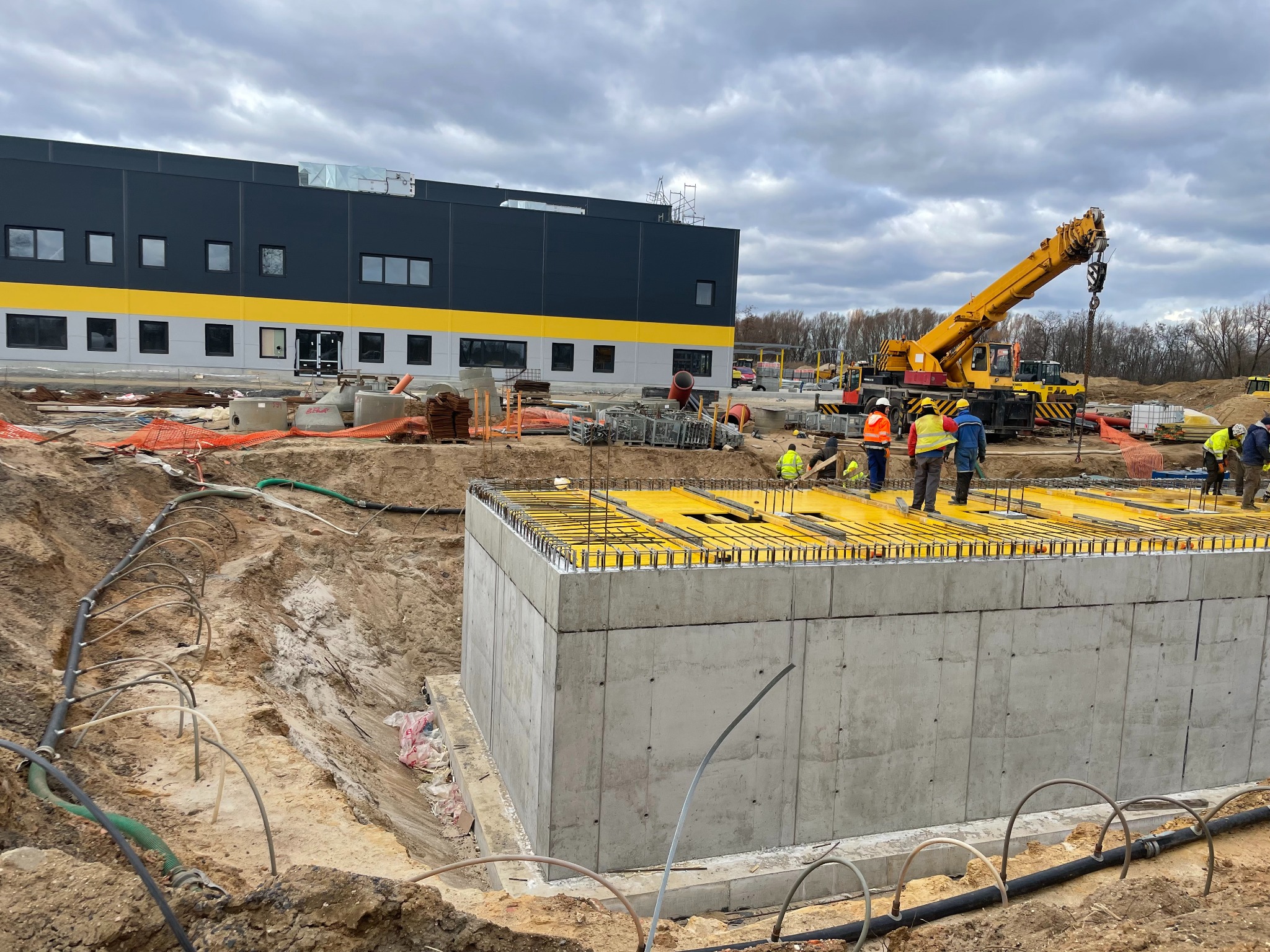 Wykop pod fundamenty z betonową konstrukcją szalunkową, zbrojeniem i żółtymi panelami na górze, robotnicy w odblaskowych kamizelkach, żuraw budowlany w tle, budynek przemysłowy w tle, pochmurne niebo.