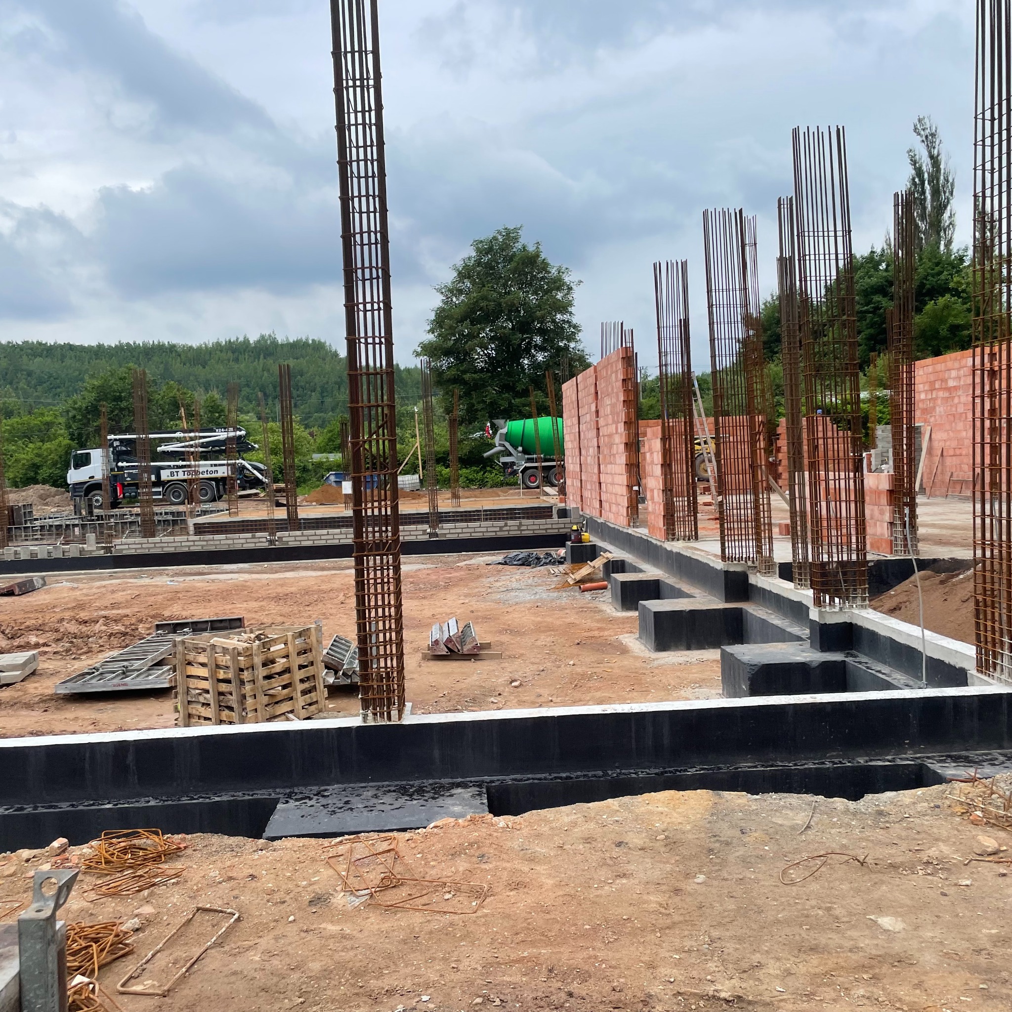 Fundamenty domu w trakcie budowy z widocznym zbrojeniem, izolacją przeciwwilgociową, częściowo wymurowanymi ścianami i betonomieszarkami w tle.