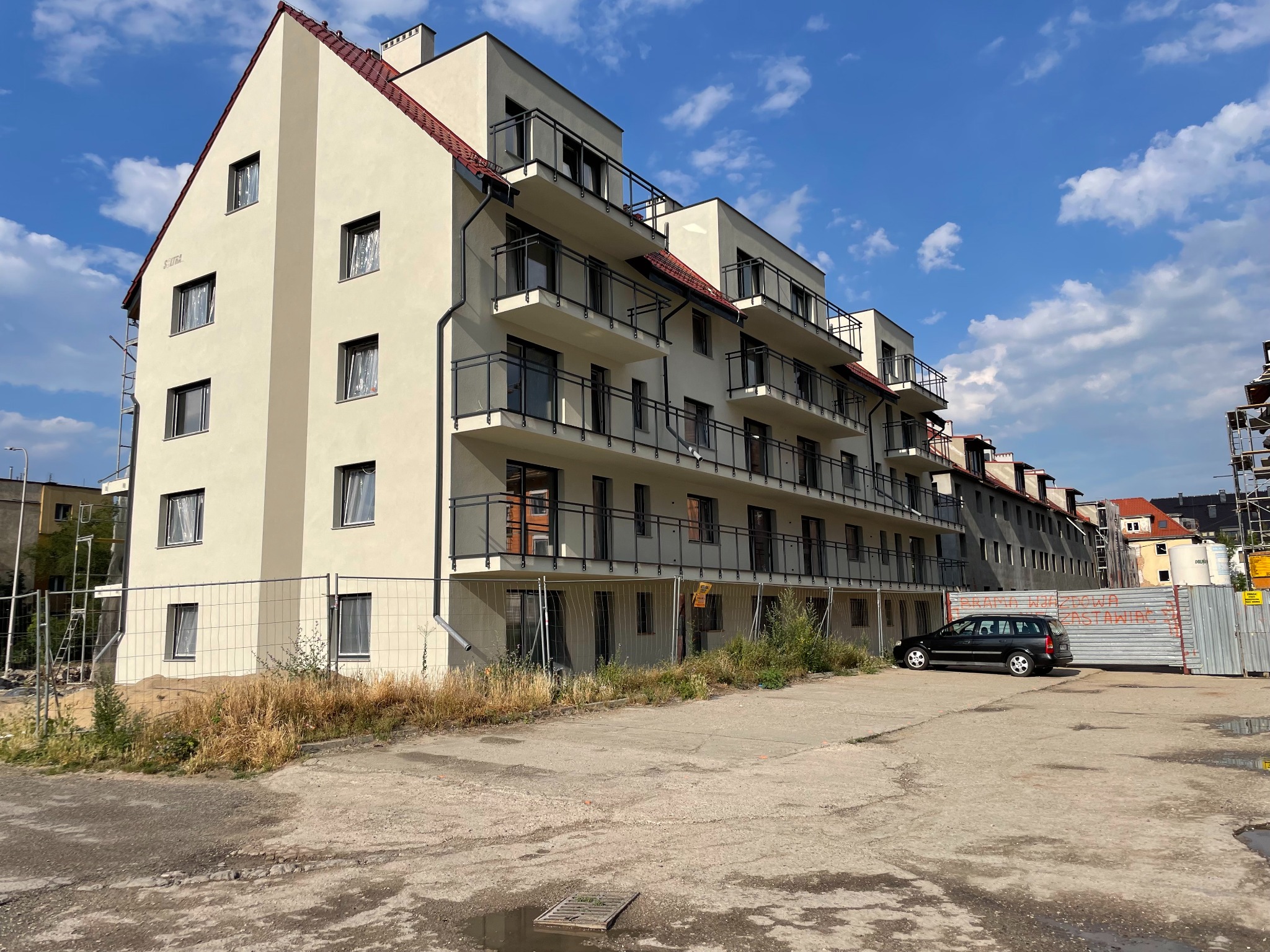 Nowoczesny, trzypiętrowy budynek mieszkalny z balkonami, w trakcie wykańczania, widoczny z zewnątrz, z zaparkowanym samochodem osobowym i ogrodzeniem placu budowy w tle.