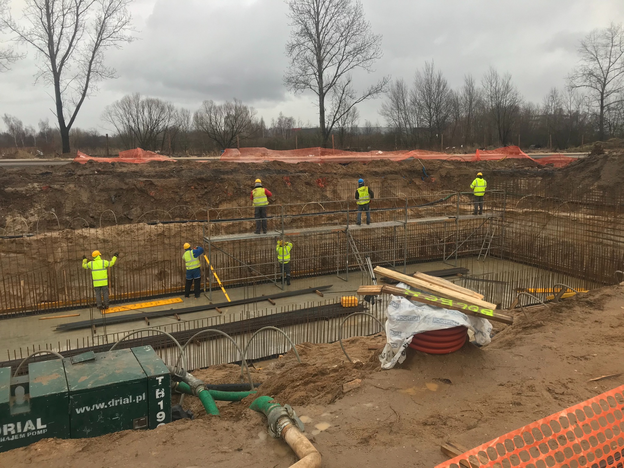 Budowa fundamentów: pracownicy w żółtych kamizelkach i kaskach na rusztowaniu, zbrojenie, wylany beton, pompa odwadniająca z zielonymi wężami, ziemia i drzewa w tle pod pochmurnym niebem.