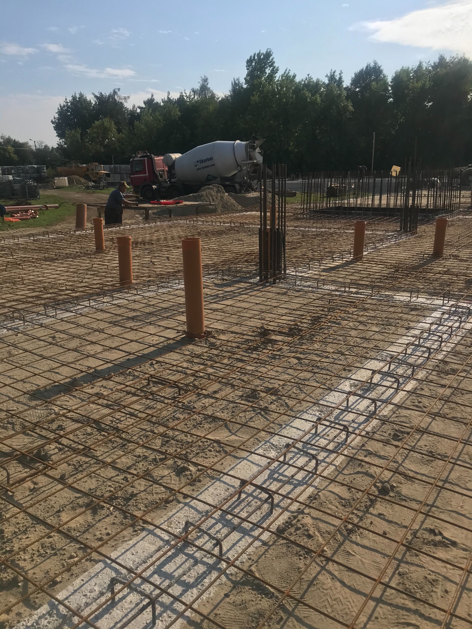 Zbrojenie fundamentów z pionowymi rurami kanalizacyjnymi, w tle betoniarka i pracownik budowlany.