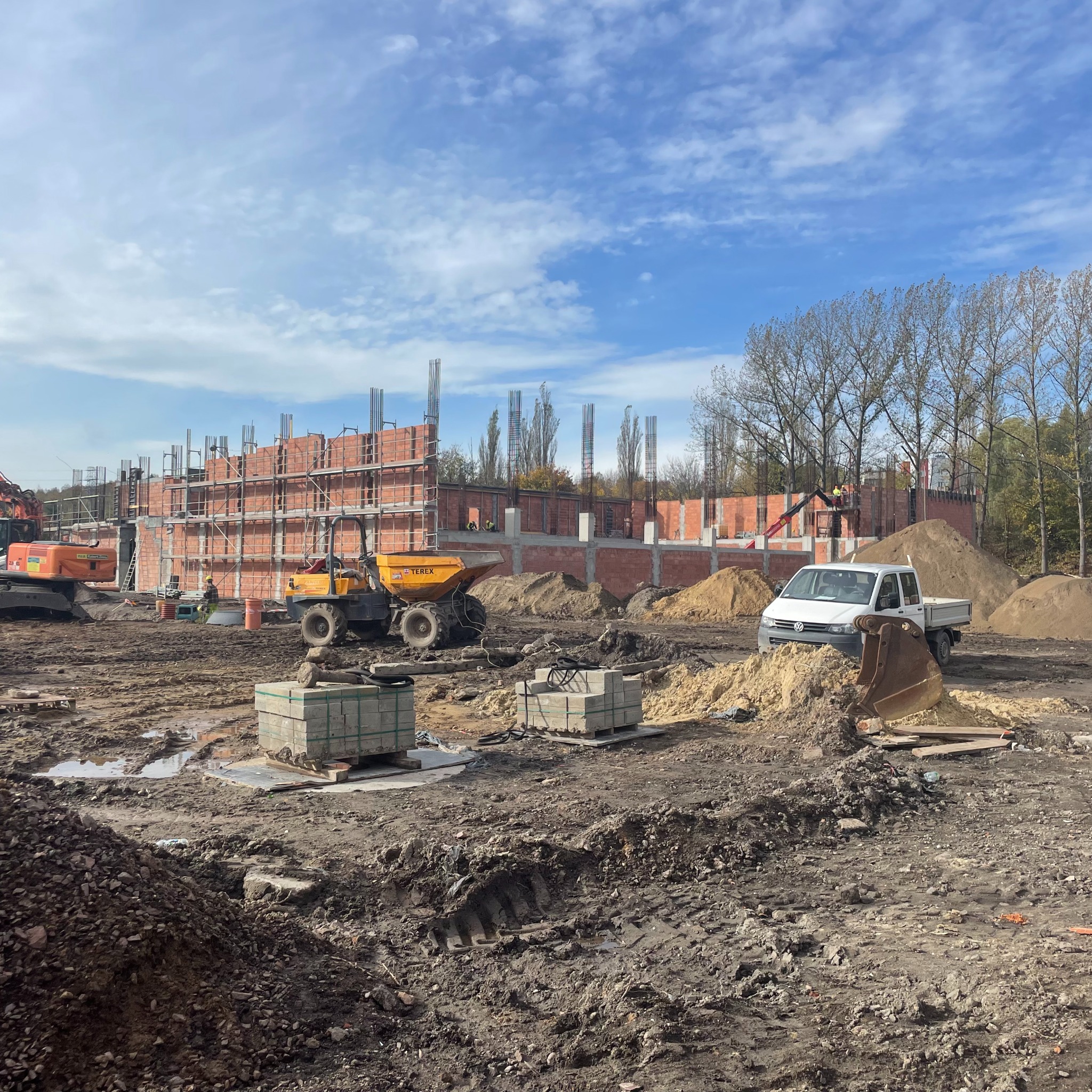 Budowa domu z czerwonej cegły w trakcie realizacji, widoczne maszyny budowlane: koparka, wywrotka, oraz stosy cegieł i piasku na placu budowy pod błękitnym niebem.