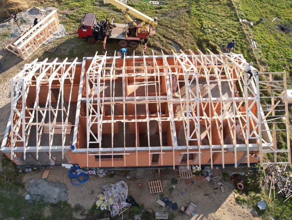 Widok z drona na budowę domu murowanego z drewnianą konstrukcją dachu w trakcie montażu, z widocznym dźwigiem i pracownikami na placu budowy.