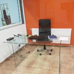 Biuro z pomarańczową szklaną ścianą, wyposażone w przezroczyste szklane biurko z półką, czarne krzesło biurowe i biały grzejnik.