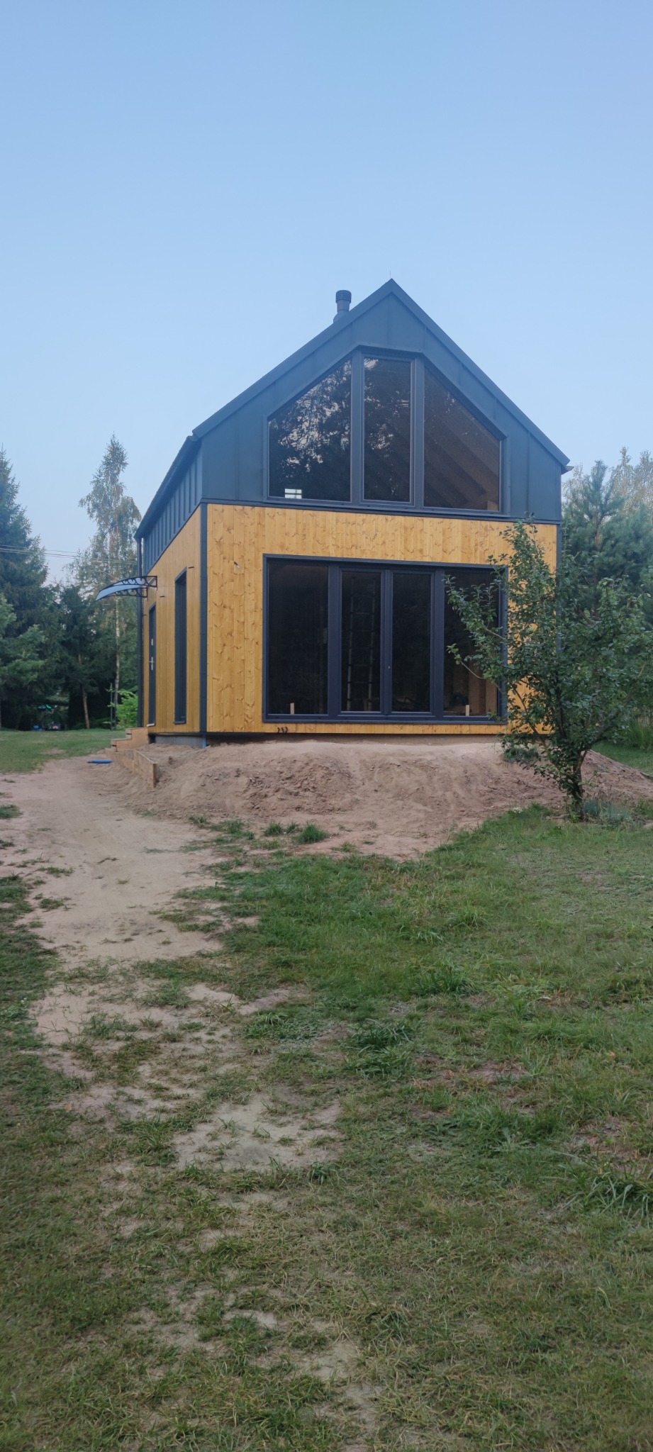 Nowoczesny dom z drewnianą elewacją i dużymi oknami, otoczony zielenią. Widoczne elementy wykończeniowe i minimalistyczny design.