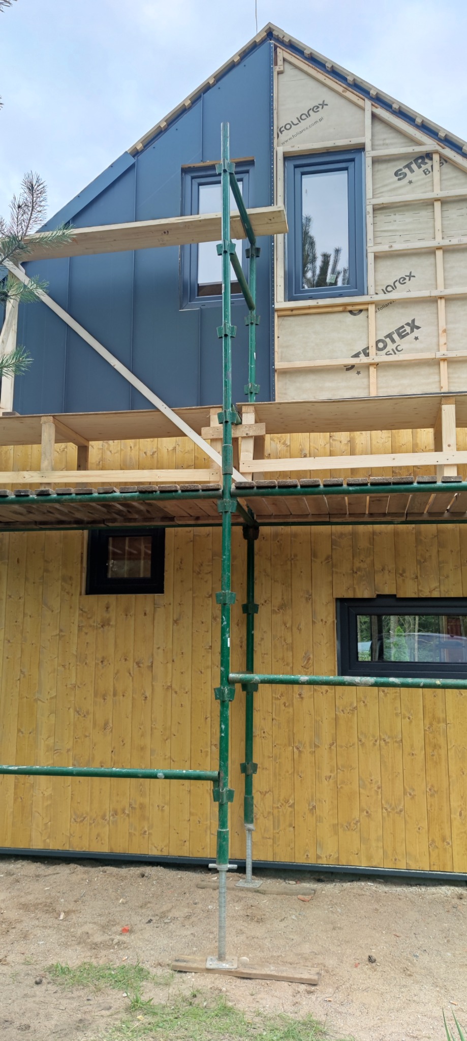 Dom w trakcie wykończenia elewacji: część pokryta szarą blachą, część ocieplona, okna wstawione, rusztowanie. Widok z dołu, ukazujący postęp prac.