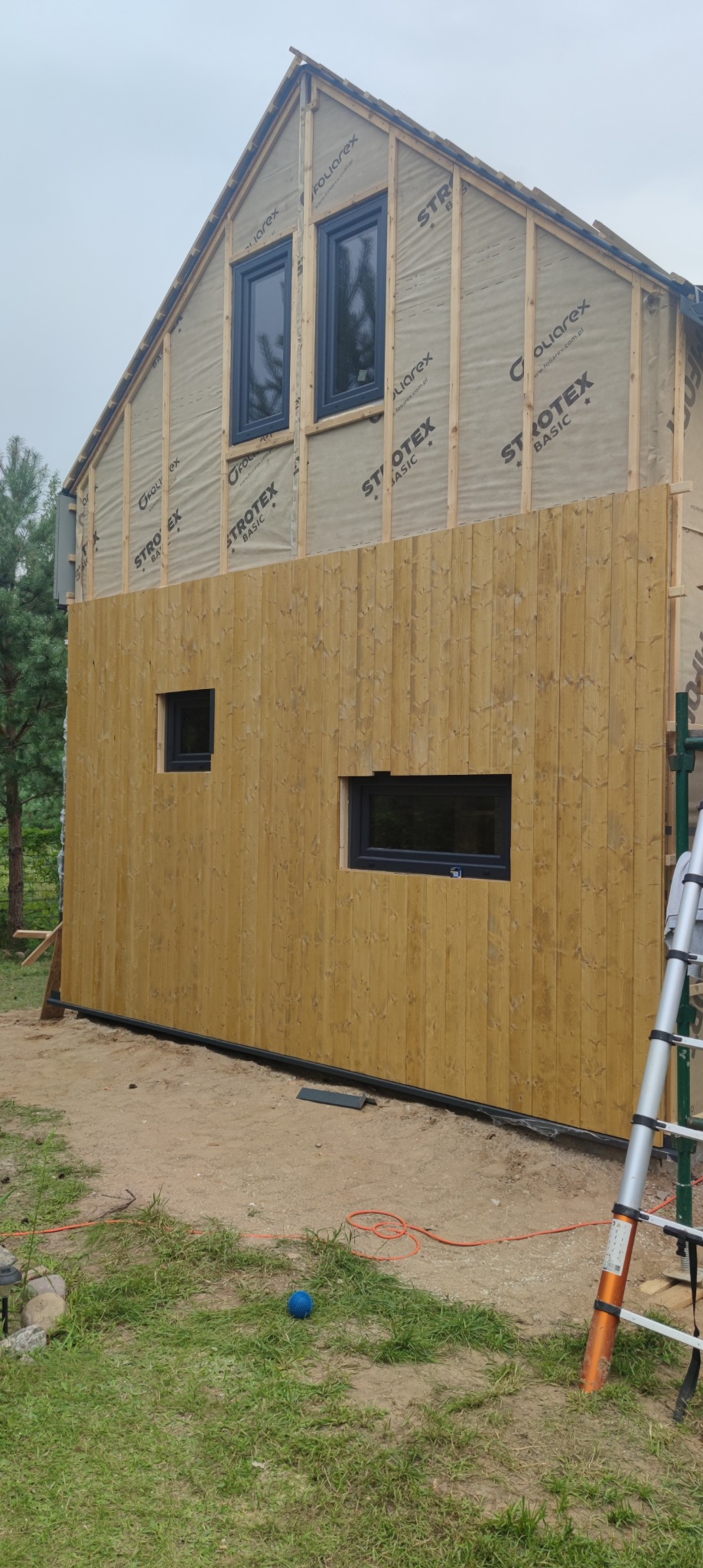 Dom w trakcie budowy z widoczną drewnianą elewacją na dolnej połowie i folią izolacyjną na górnej. Okna wstawione, drabina oparta o ścianę. Częściowo zakończony etap prac.