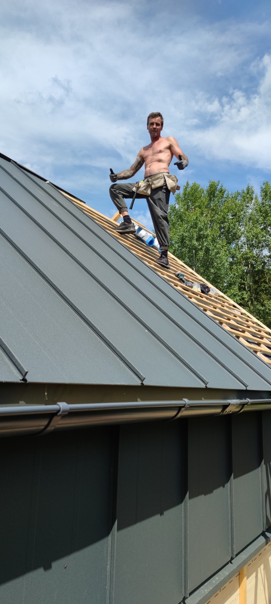 Dekarz bez koszulki z tatuażem na dachu z szarej blachy trapezowej, w pasie narzędziowym i rękawicach, kciuki w górę, w tle drzewa i błękitne niebo.