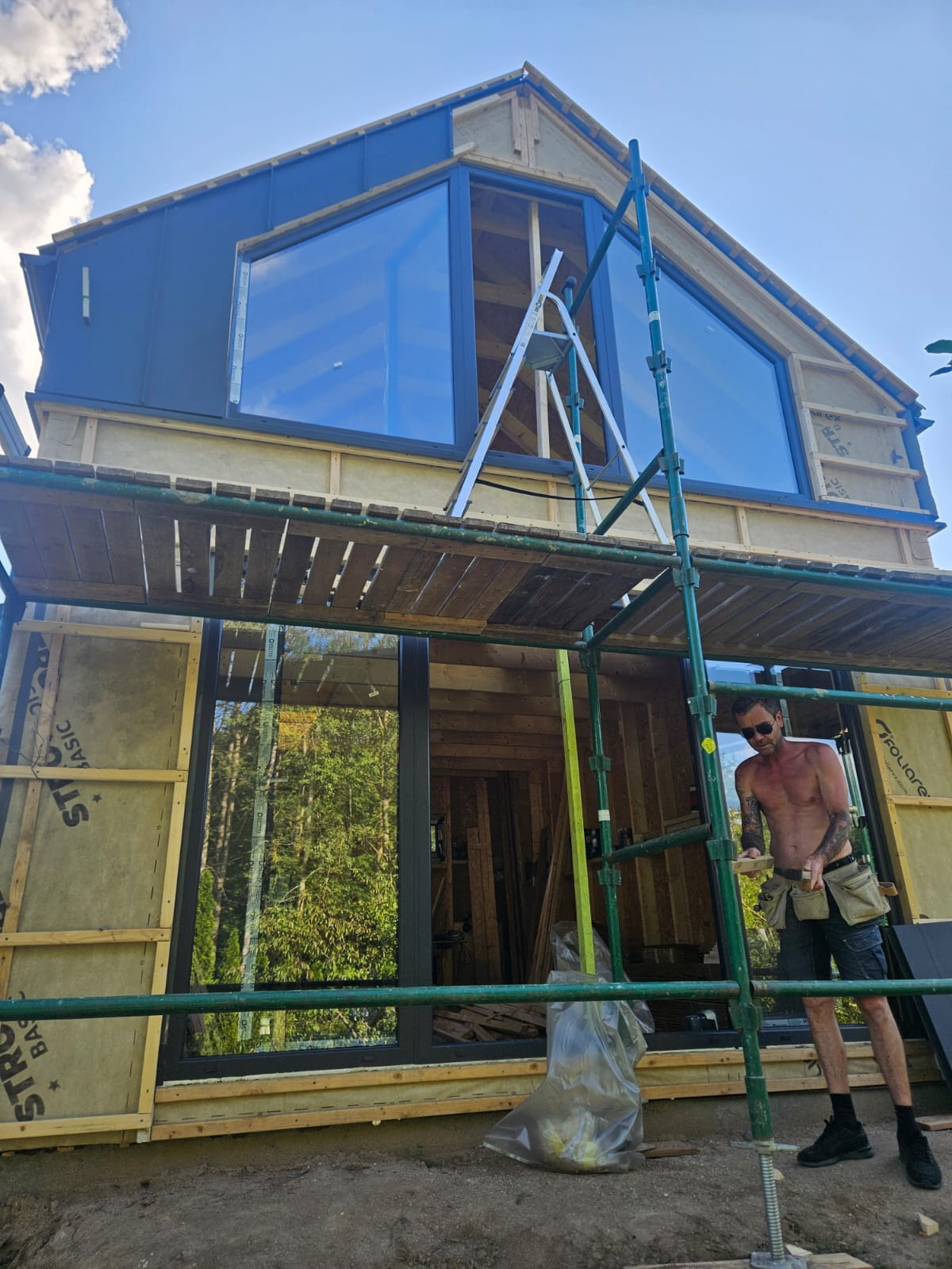 Budowa domu z dużymi oknami, widok z dołu na rusztowanie i pracownika w trakcie montażu, w tle las i błękitne niebo. Dom w stanie surowym otwartym.