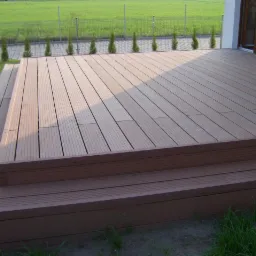 Nowoczesny taras z kompozytowych desek w odcieniach brązu, z dwustopniowym wejściem, otoczony zielonym trawnikiem i tujami, widok na ogrodzenie i pole w tle.