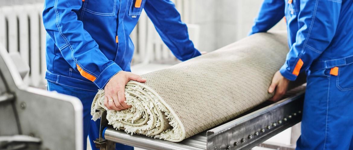 Dwaj pracownicy w niebieskich kombinezonach przenoszą zwinięty beżowy dywan z frędzlami na metalowej konstrukcji.