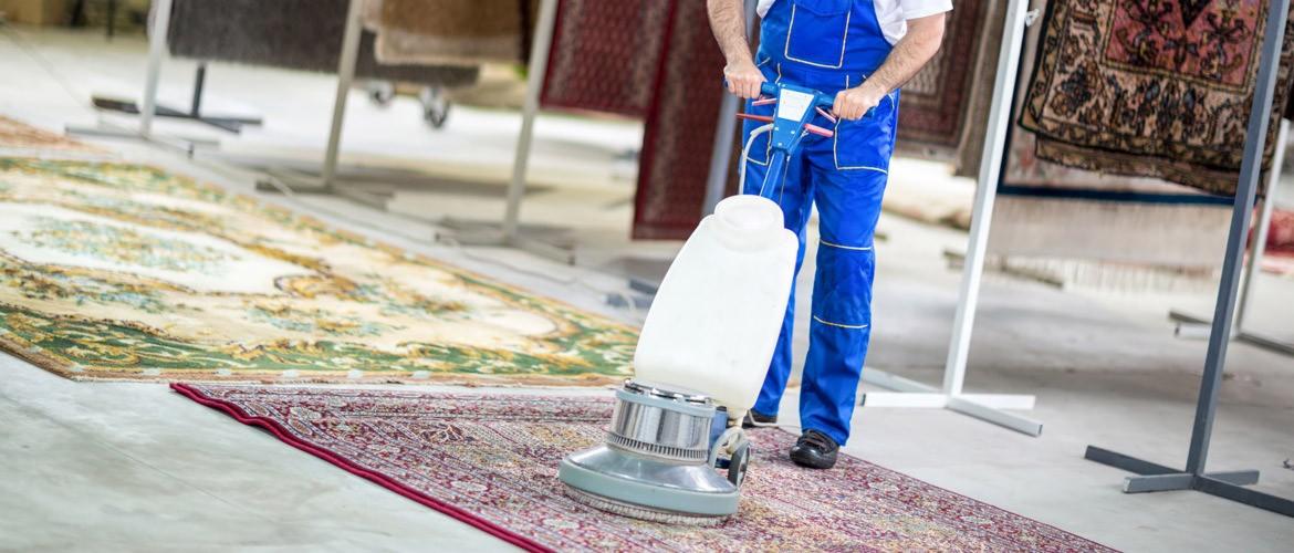 Pracownik w niebieskim kombinezonie czyści dywan orientalny za pomocą profesjonalnej maszyny czyszczącej w jasnym pomieszczeniu z dywanami na stojakach w tle.